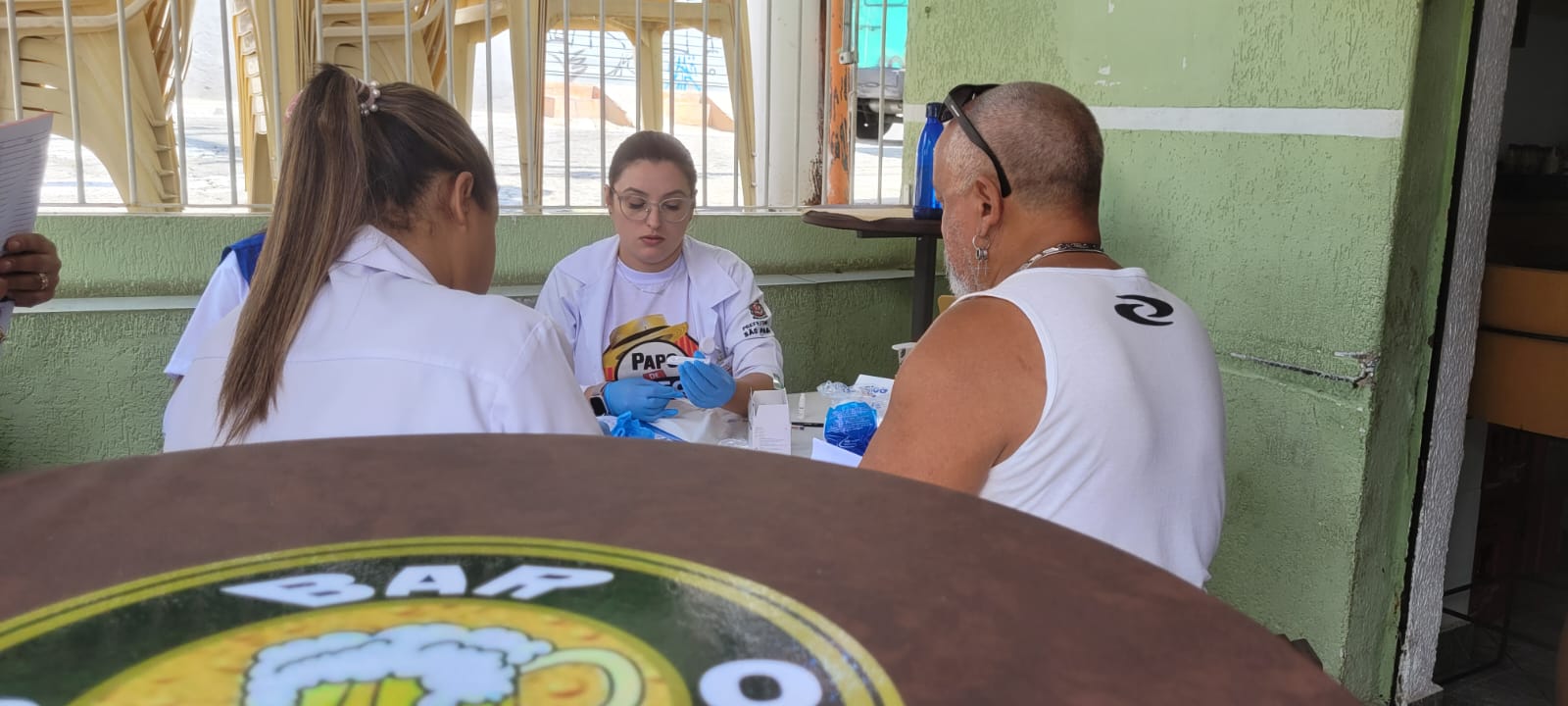 Na imagem, uma equipe da UBS atende um idoso que participa da ação. Ele está de costas, é careca, usa regata branca, brinco, corrente prateada e está um óculos de sol  na cabeça